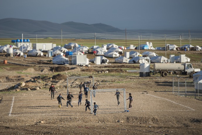 Children playing soccer on a dirt field beside a refugee camp